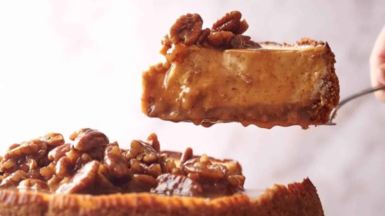 A slice of pecan-topped cheesecake is held above the rest of the cake.