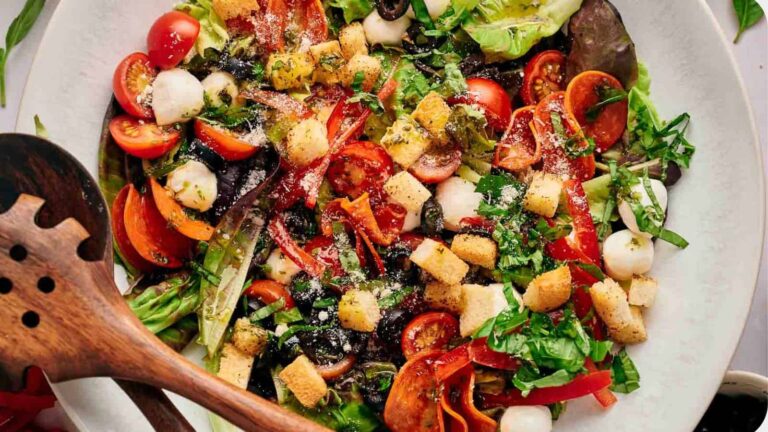 A colorful salad with mixed greens, cherry tomatoes, mozzarella balls, croutons, and sliced red peppers, topped with herbs and a sprinkling of grated cheese. Wooden salad utensils on the side.