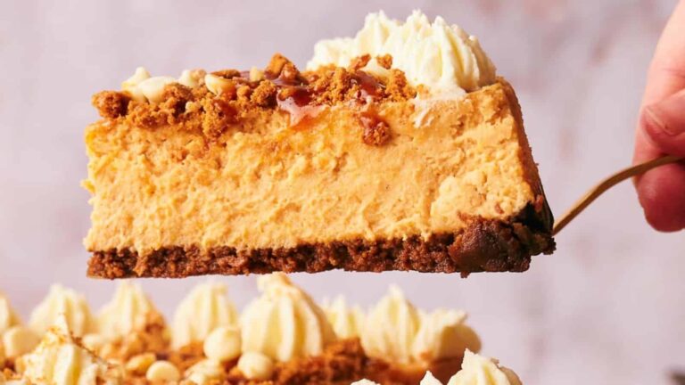 A slice of creamy cheesecake with a crumbly base, topped with whipped cream, crumbled nuts, and caramel drizzle.