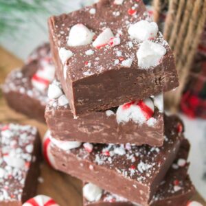 A stack of peppermint fudge squares, topped with crushed candy canes, sits on a wooden board with whole peppermint candies nearby.