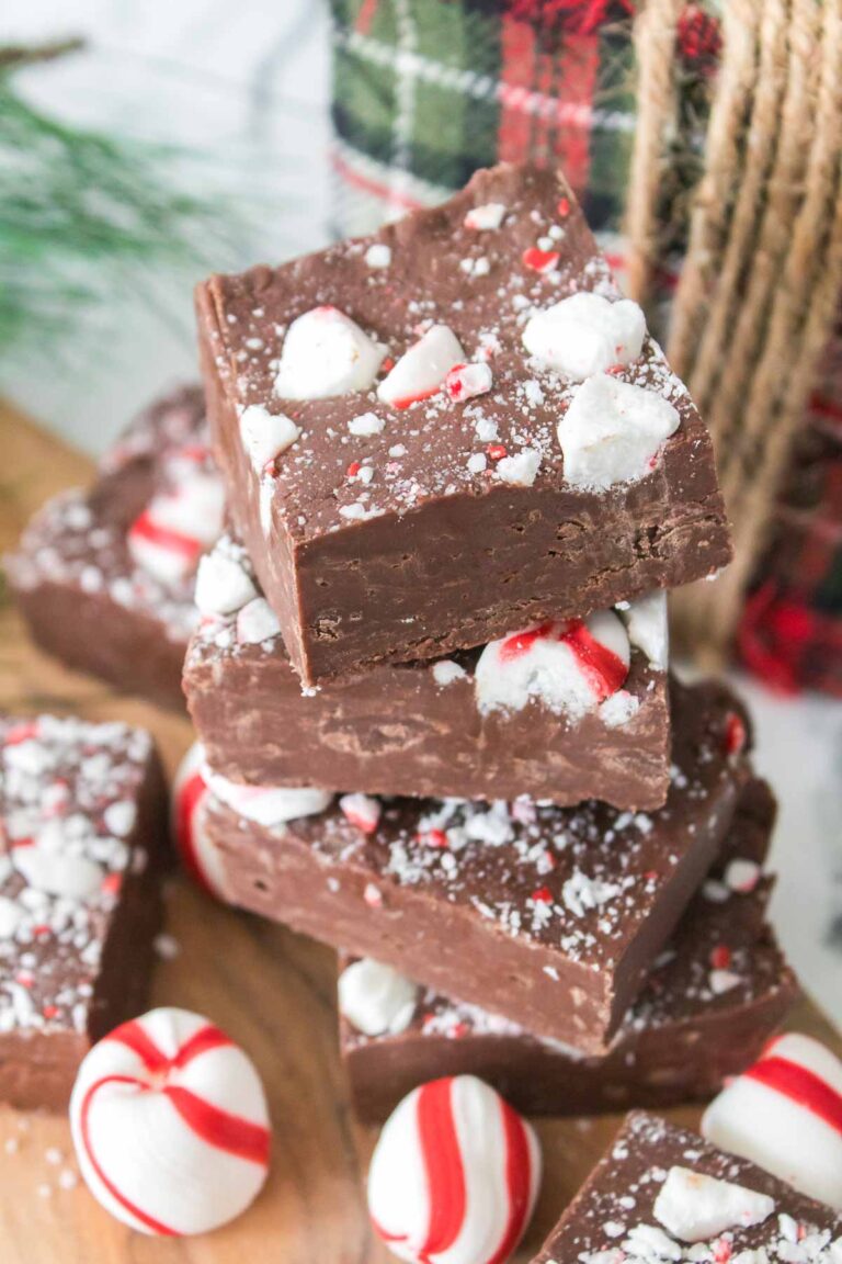 A stack of peppermint fudge squares, topped with crushed candy canes, sits on a wooden board with whole peppermint candies nearby.