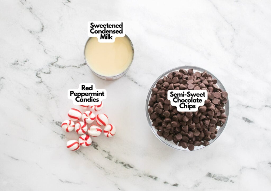 A can of sweetened condensed milk, a bowl of semi-sweet chocolate chips, and a small pile of red peppermint candies on a marble surface&mdash;perfect ingredients for creamy peppermint fudge.