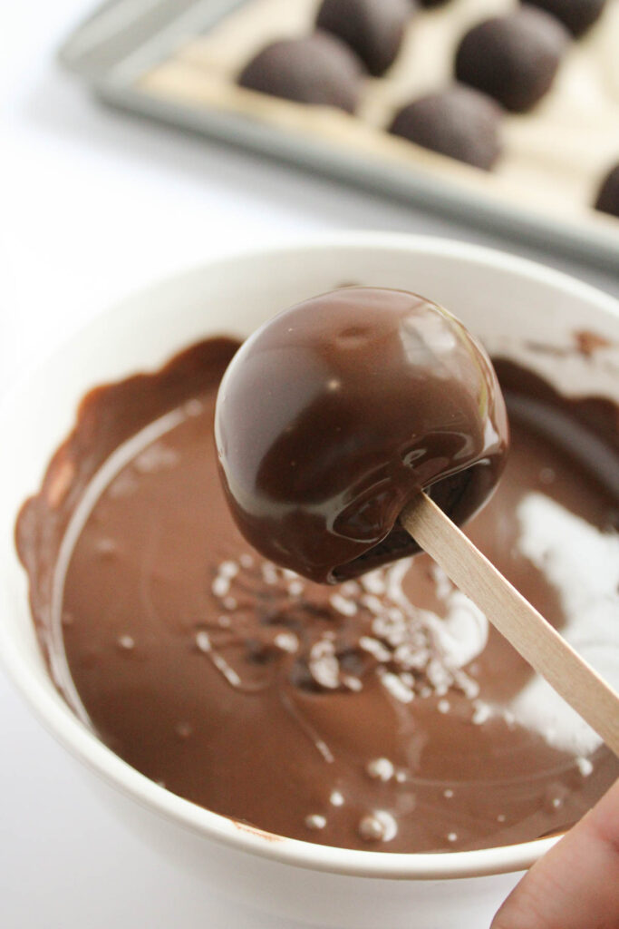 A festive reindeer cake pop is being dipped into melted chocolate in a white bowl, creating a delightful holiday treat.