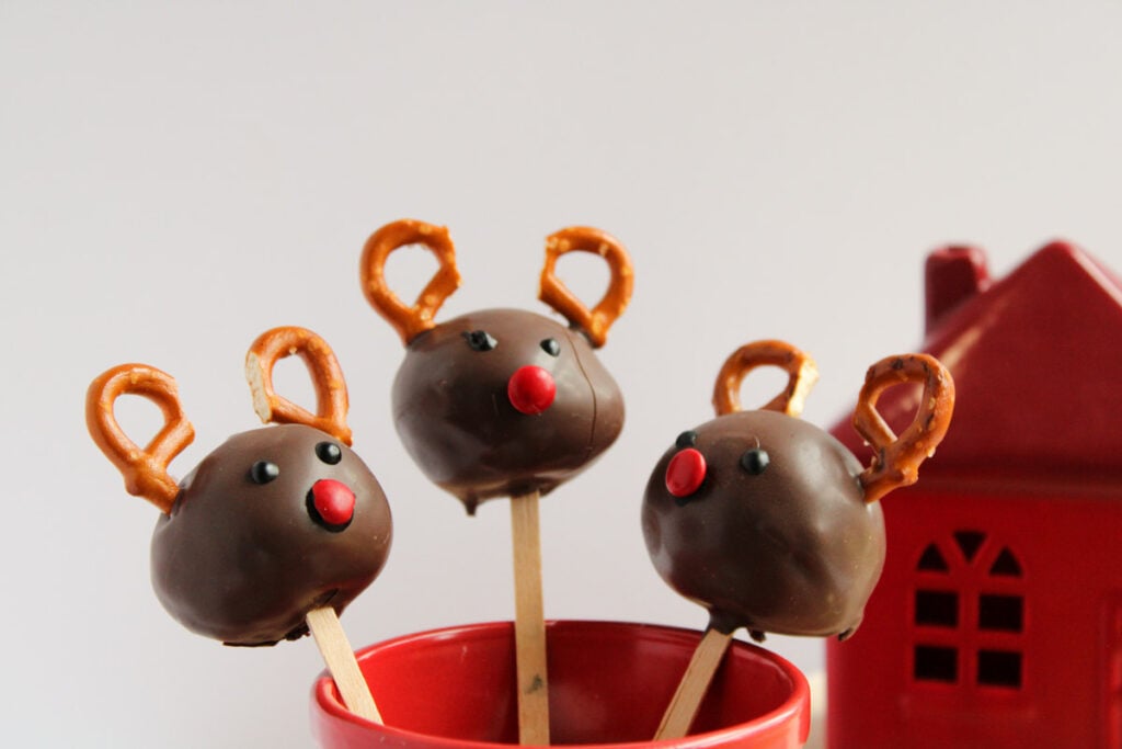Three Reindeer Cake Pops decorated with pretzel antlers and red candy noses are placed in a red cup next to a small red house decoration.