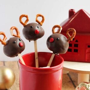 Three reindeer cake pops with chocolate coating, pretzel antlers, and red candy noses stand in a red container, beside a red decorative house and ornaments.