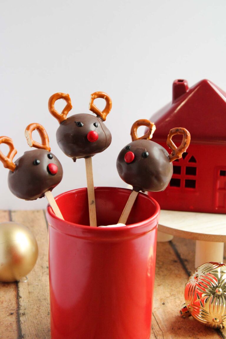 Three reindeer cake pops with chocolate coating, pretzel antlers, and red candy noses stand in a red container, beside a red decorative house and ornaments.