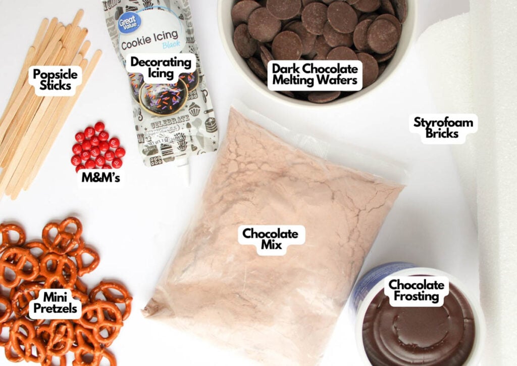 Various ingredients and supplies for baking Reindeer Cake Pops, including popsicle sticks, decorating icing, M&M’s, mini pretzels, chocolate mix, dark chocolate wafers, styrofoam bricks, and chocolate frosting.