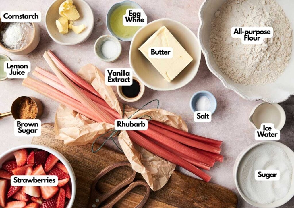Top-down view of baking ingredients for a classic rhubarb pie, featuring strawberries, butter, sugar, flour, egg white, cornstarch, vanilla extract, lemon juice, brown sugar, salt, and ice water.