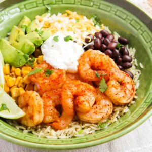 A green bowl with shrimp, rice, avocado, corn, black beans, cheese, sour cream, and lime wedge, garnished with cilantro.