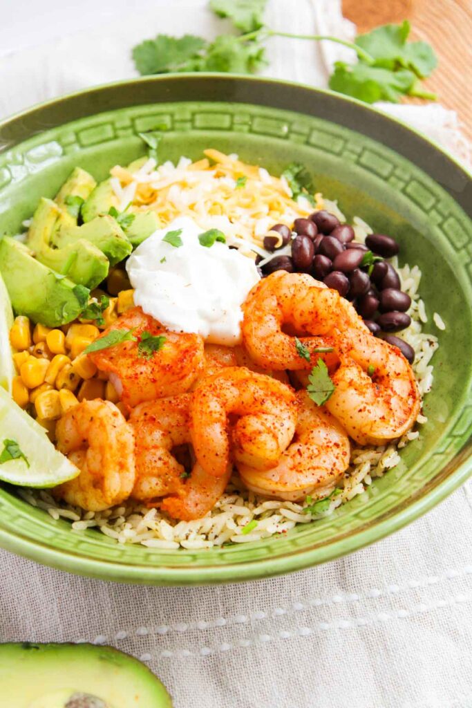 A green bowl with shrimp, rice, avocado, corn, black beans, cheese, sour cream, and lime wedge, garnished with cilantro.