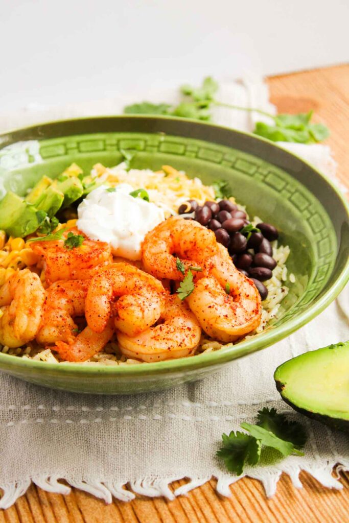 A green bowl filled with seasoned shrimp, diced avocado, corn, black beans, rice, shredded cheese, and a dollop of sour cream, garnished with cilantro on a wooden surface.