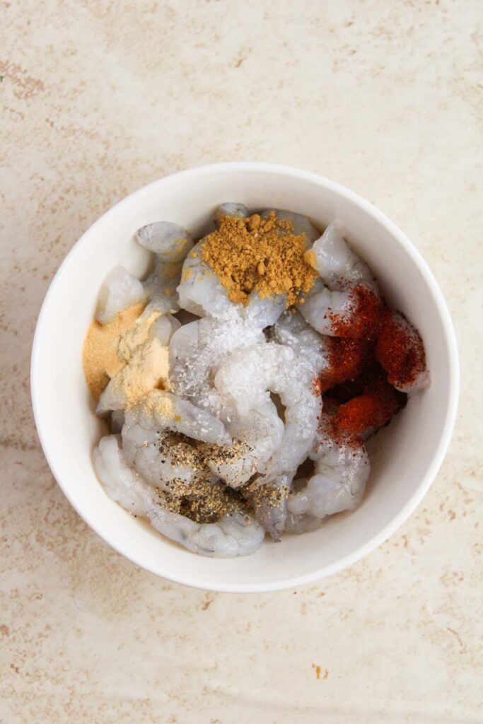 A white bowl containing raw shrimp with various spices, including paprika, salt, and black pepper, on a beige surface.