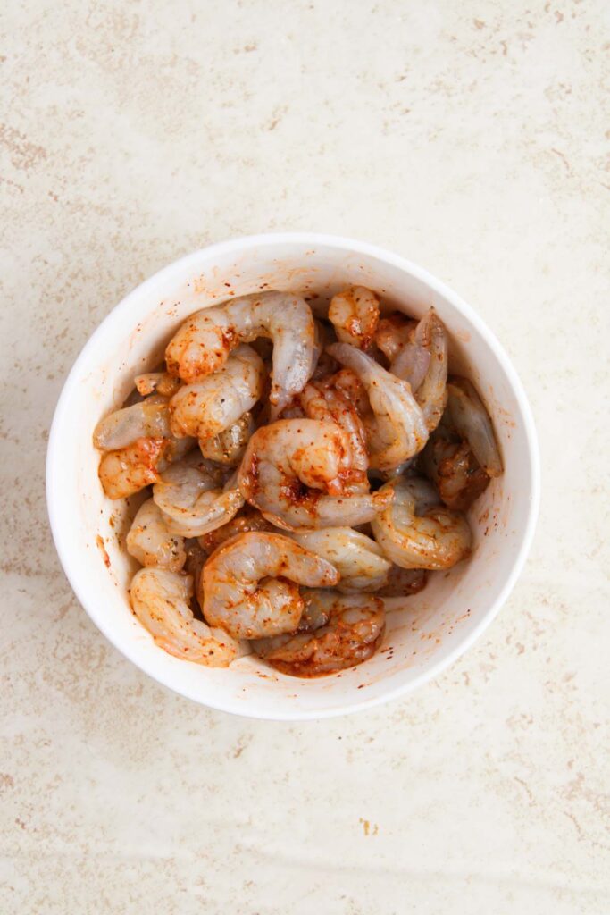 A bowl of raw shrimp marinated with spices sits on a light-colored surface.