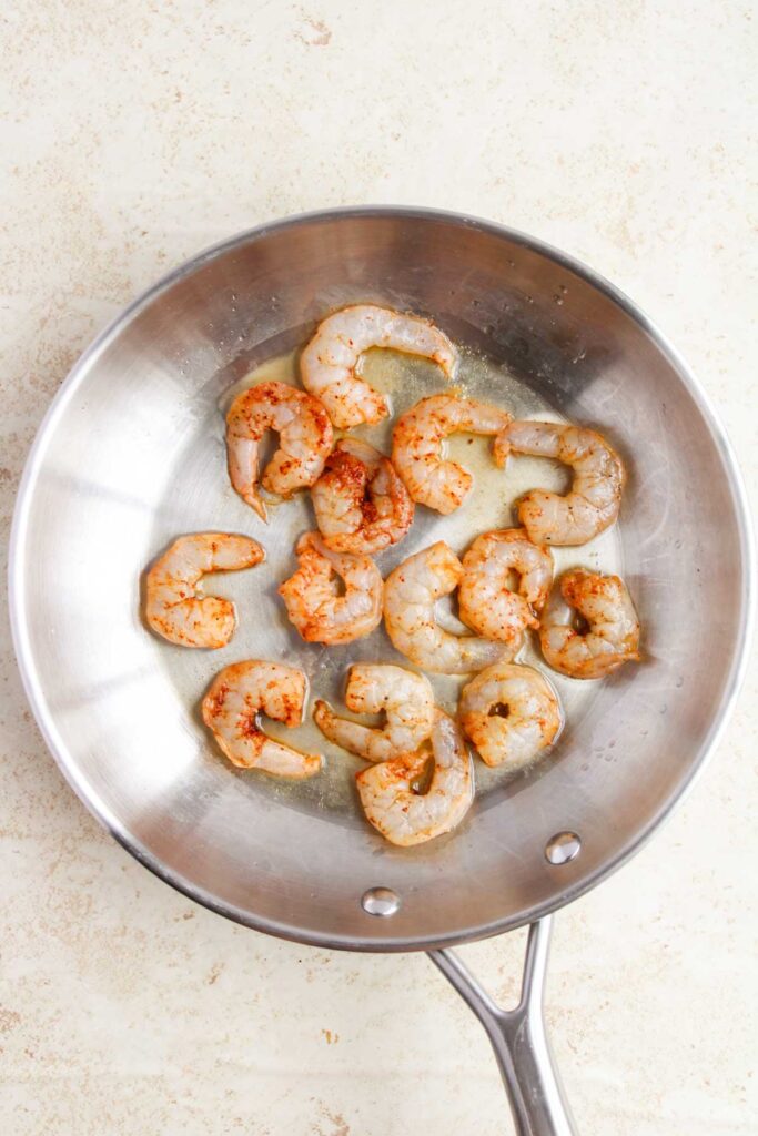 A stainless steel pan containing raw shrimp seasoned with spices.