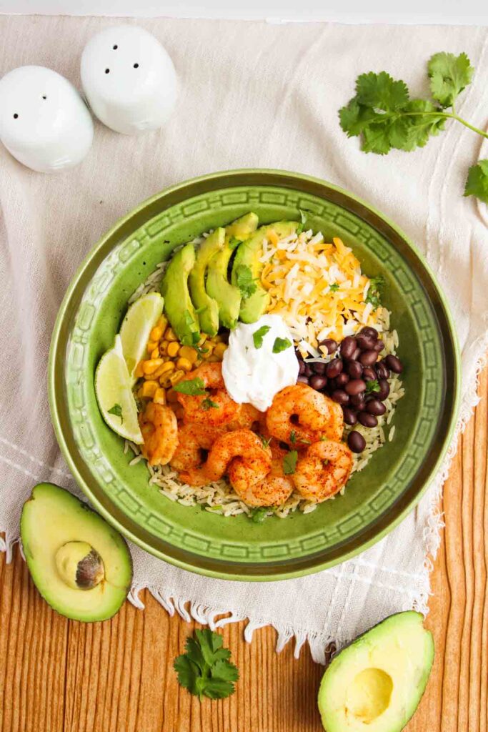 A green bowl with shrimp, avocado slices, corn, black beans, rice, grated cheese, and a dollop of sour cream. Lime wedges are on the side. Avocado halves and cilantro garnish the scene.
