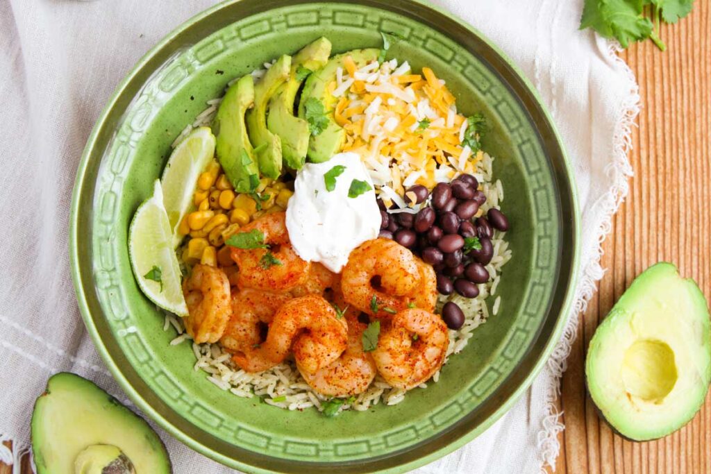 A bowl with shrimp, black beans, corn, avocado slices, shredded cheese, rice, and sour cream, garnished with lime wedges. Avocado halves on the side.