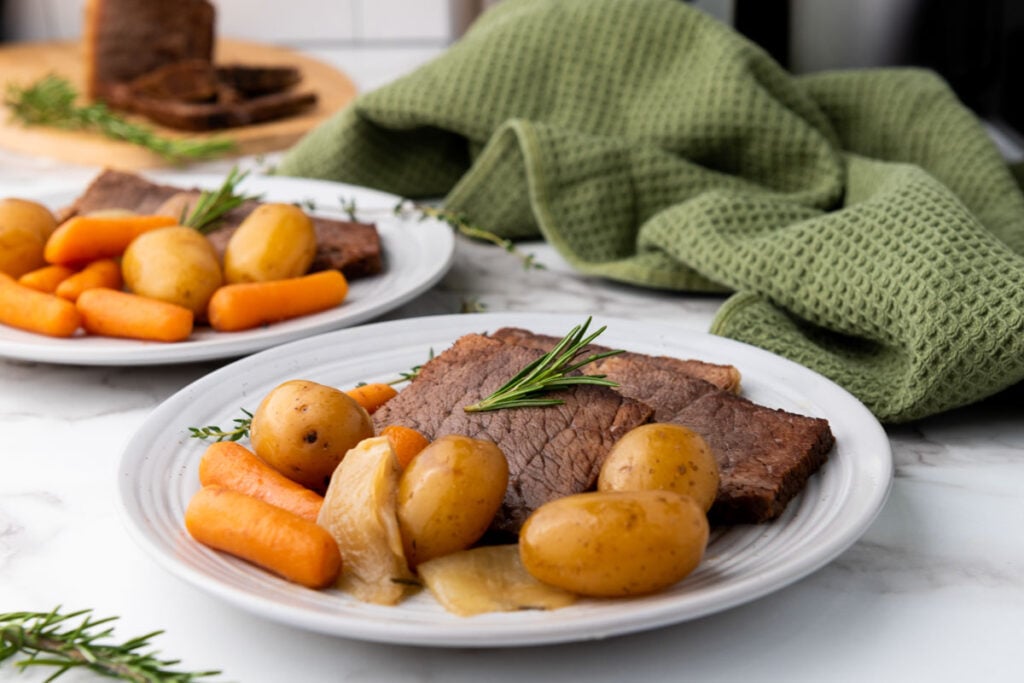 A plate with slices of roast beef, baby potatoes, carrots, onion, and a sprig of rosemary, with a green towel in the background.
