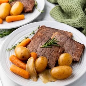 Two plates with sliced roast beef, baby carrots, potatoes, and onions, garnished with rosemary. A green cloth is in the background.