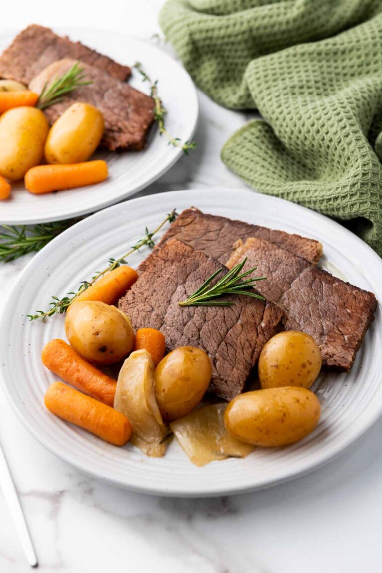 Two plates with sliced roast beef, baby carrots, potatoes, and onions, garnished with rosemary. A green cloth is in the background.