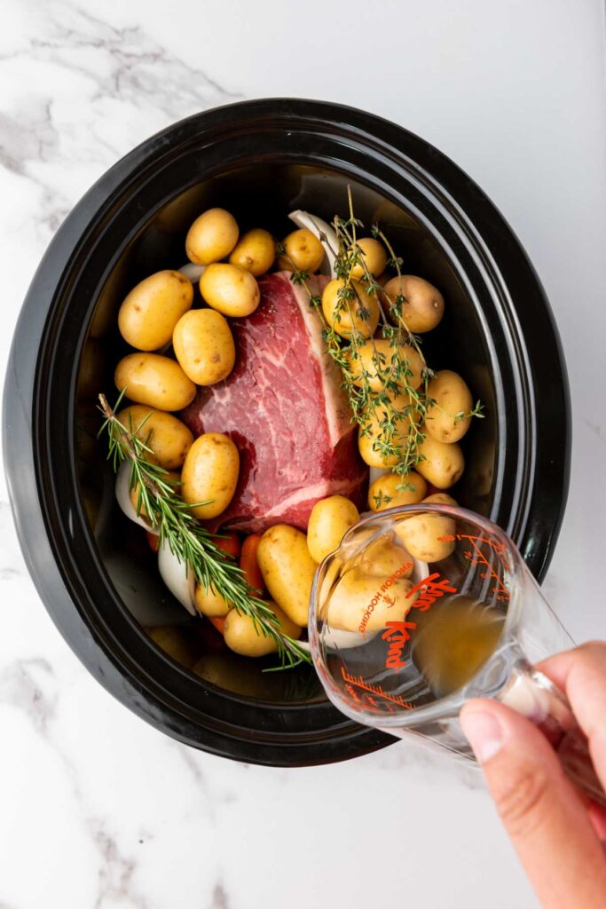 Slow cooker with beef, baby potatoes, onions, rosemary, and thyme, on white marble. A hand pours broth from a measuring cup into the pot.