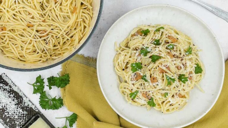 A bowl of spaghetti aglio e olio garnished with parsley and cheese, next to a pan of spaghetti, a grater, and more parsley on a yellow cloth.