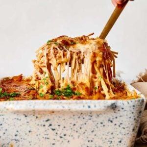 A person lifts a portion of cheesy baked pasta from a speckled ceramic dish with a fork, displaying melted cheese strands.