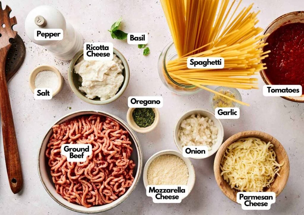 Ingredients for a Baked Spaghetti Casserole are arranged on a table, featuring ground beef, spaghetti, cheeses, tomato sauce, onion, garlic, seasonings, salt, and pepper. Each item is clearly labeled for easy preparation.