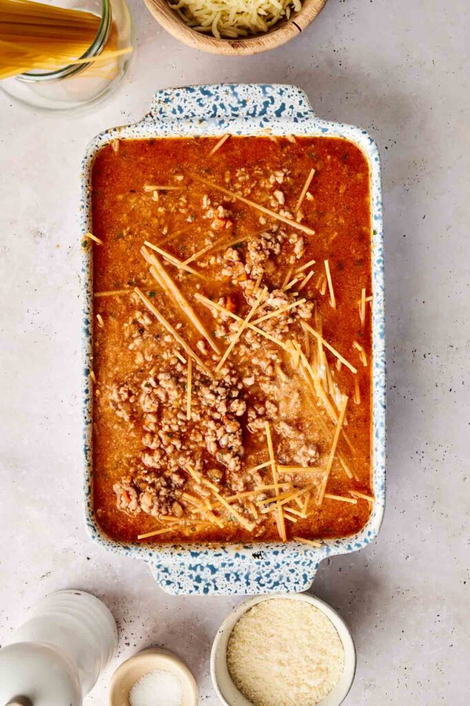 A casserole dish with layers of broken spaghetti and a meaty tomato sauce on a light surface, surrounded by ingredients like cheese and spices.