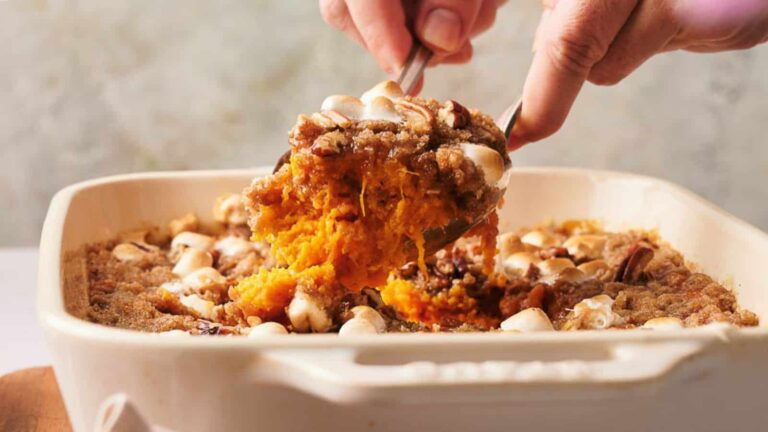 Person serving a portion of sweet potato casserole with marshmallows and pecans from a rectangular dish.
