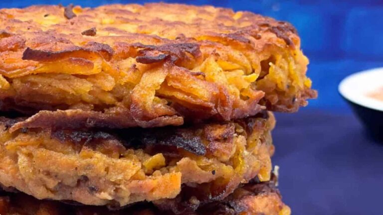 Close-up of a stack of crispy golden-brown vegetable fritters against a dark blue background, with a small bowl of dipping sauce in the distance.