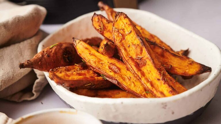 A white dish contains roasted sweet potato wedges seasoned with herbs. A beige cloth is visible beside the dish.