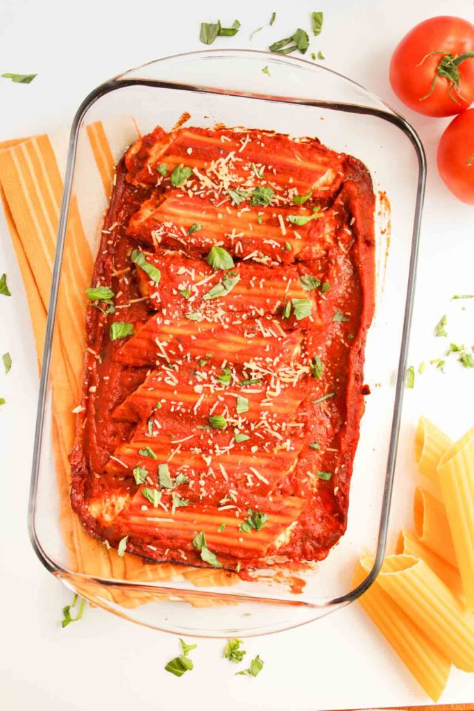 A glass baking dish brimming with Three Cheese Manicotti is topped with rich tomato sauce, grated cheese, and fresh basil. Nearby, uncooked pasta and ripe tomatoes await their turn in this mouthwatering creation.