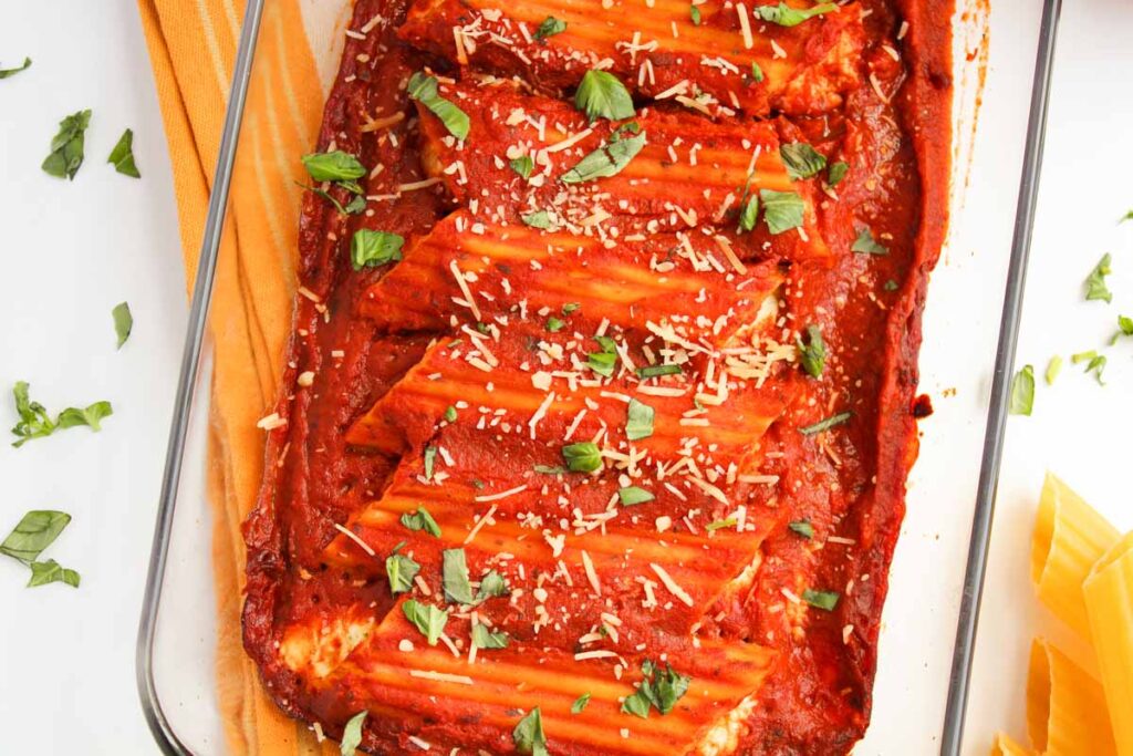 A glass baking dish holds Three Cheese Manicotti enveloped in rich red tomato sauce, sprinkled with grated cheese and adorned with fresh basil leaves.