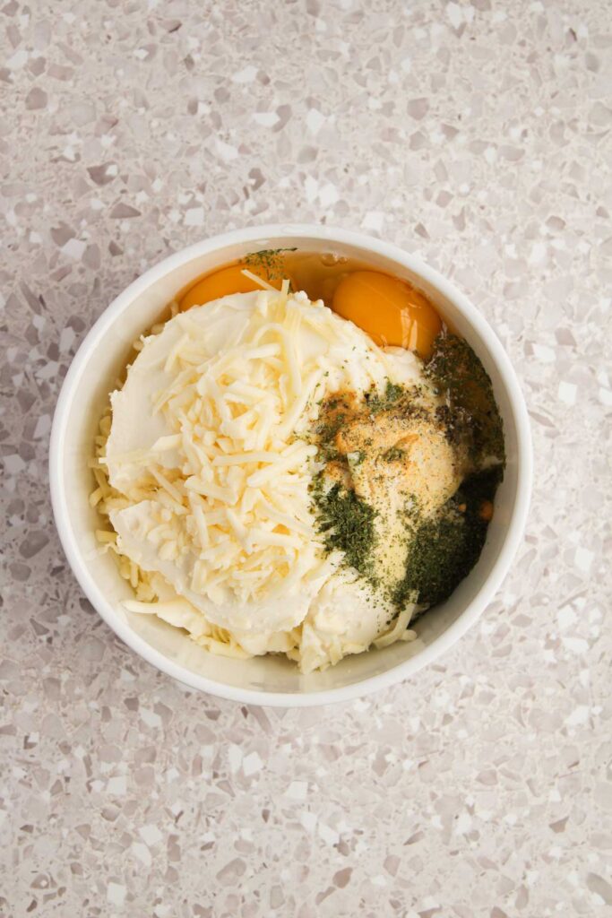 A bowl containing mashed potatoes, three cheese blend reminiscent of a manicotti filling, egg yolks, and herbs rests on a speckled countertop.