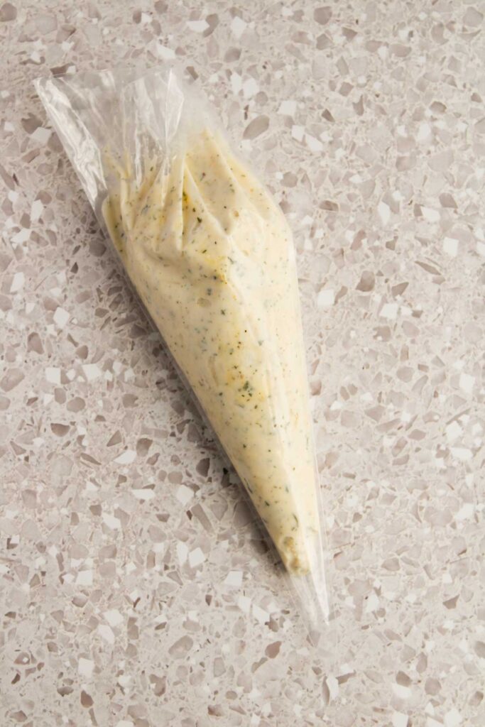 A clear piping bag filled with a creamy, herb-infused mixture, perfect for stuffing into Three Cheese Manicotti, rests on a speckled surface.