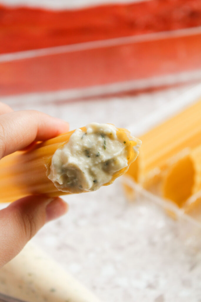 Close-up of a hand holding a large pasta shell stuffed with a creamy white filling, reminiscent of Three Cheese Manicotti, with uncooked pasta in the background.
