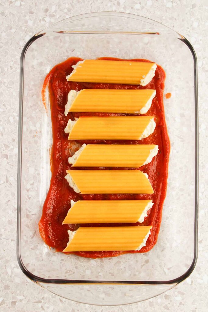 A glass baking dish with five uncooked cannelloni pasta tubes, reminiscent of Three Cheese Manicotti, filled with cheese and placed on a layer of rich tomato sauce.