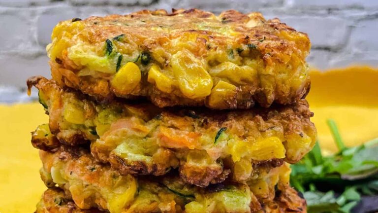 A stack of three golden-brown vegetable fritters with visible corn, zucchini, and carrot pieces. A small pile of greens is partially visible in the background.