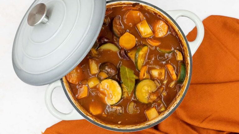 A pot of vegetable stew with carrots, zucchini, potatoes, and bay leaves, partially covered by a lid, placed on an orange cloth.
