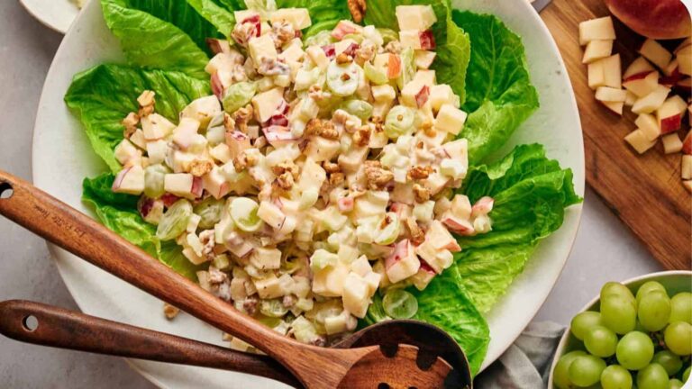 Plate of Waldorf salad with chopped apples, grapes, walnuts, and celery on lettuce leaves. Wooden serving spoon and fork on the side.