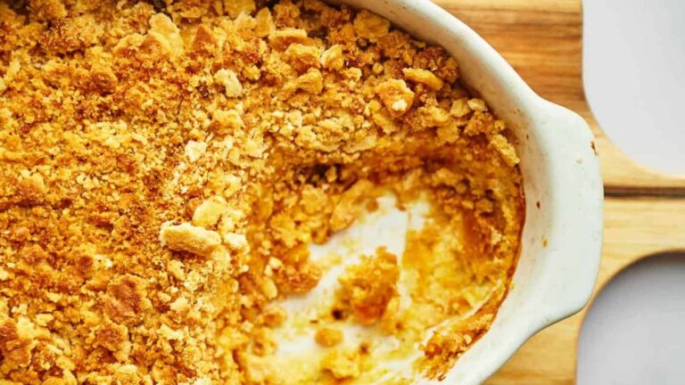 A partially eaten dish of golden, crumbly casserole sits in a white ceramic baking dish on a wooden surface.