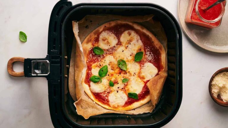 A pizza with cheese and basil on parchment paper in an air fryer. A small bowl of grated cheese and a jar of sauce are nearby.