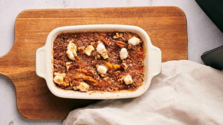 A baked casserole in a rectangular dish on a wooden board, topped with pecans and dollops of soft cheese. A beige cloth is partially visible to the right.