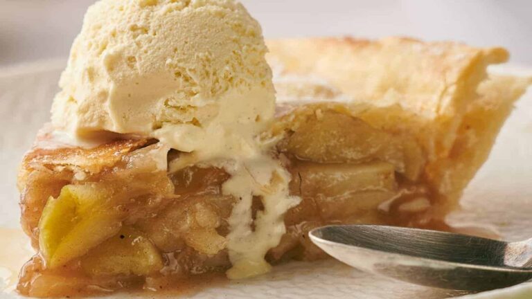 Slice of apple pie topped with a scoop of vanilla ice cream, next to a spoon.