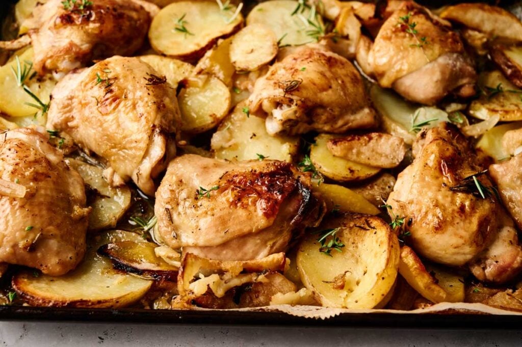 Baked chicken thighs and sliced potatoes with herbs, roasted together on a baking sheet.