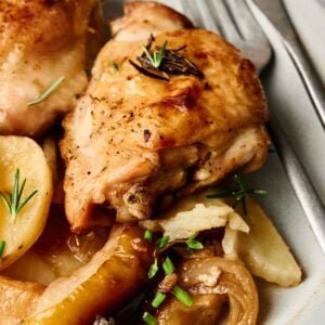 Plate with baked chicken thighs, garnished with herbs, accompanied by potato slices and vegetables. Silverware is placed beside the plate.
