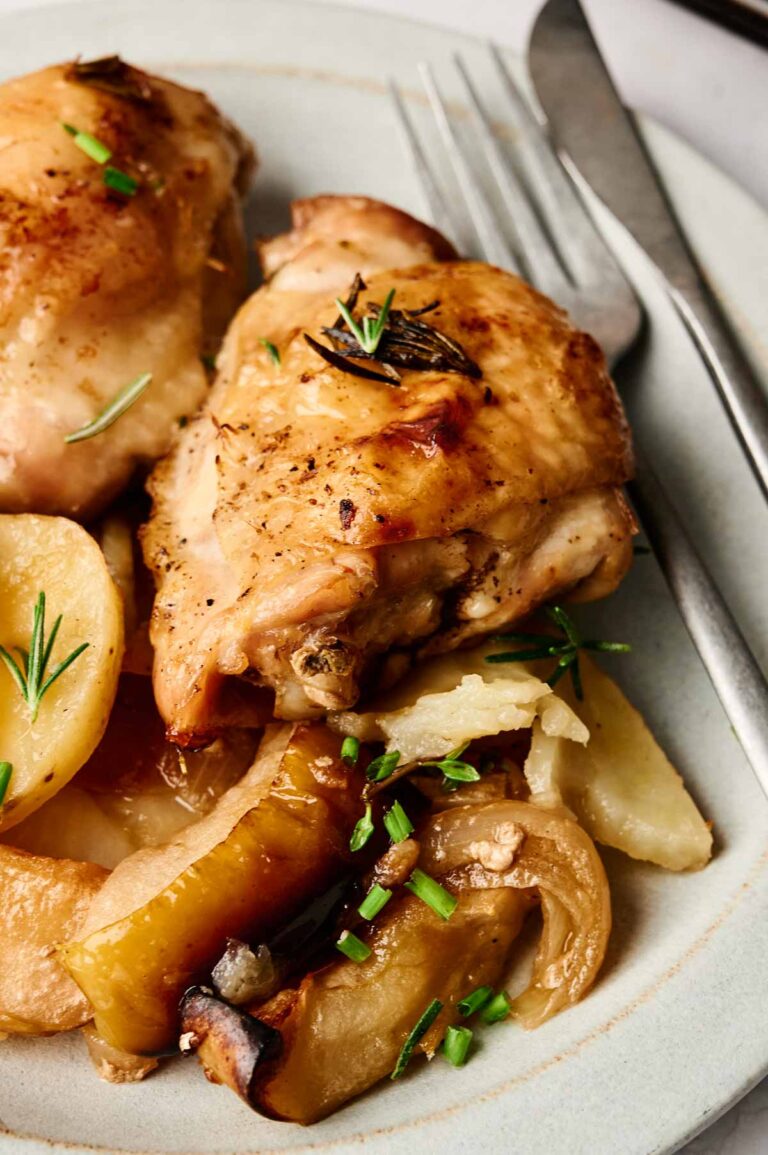 Plate with baked chicken thighs, garnished with herbs, accompanied by potato slices and vegetables. Silverware is placed beside the plate.