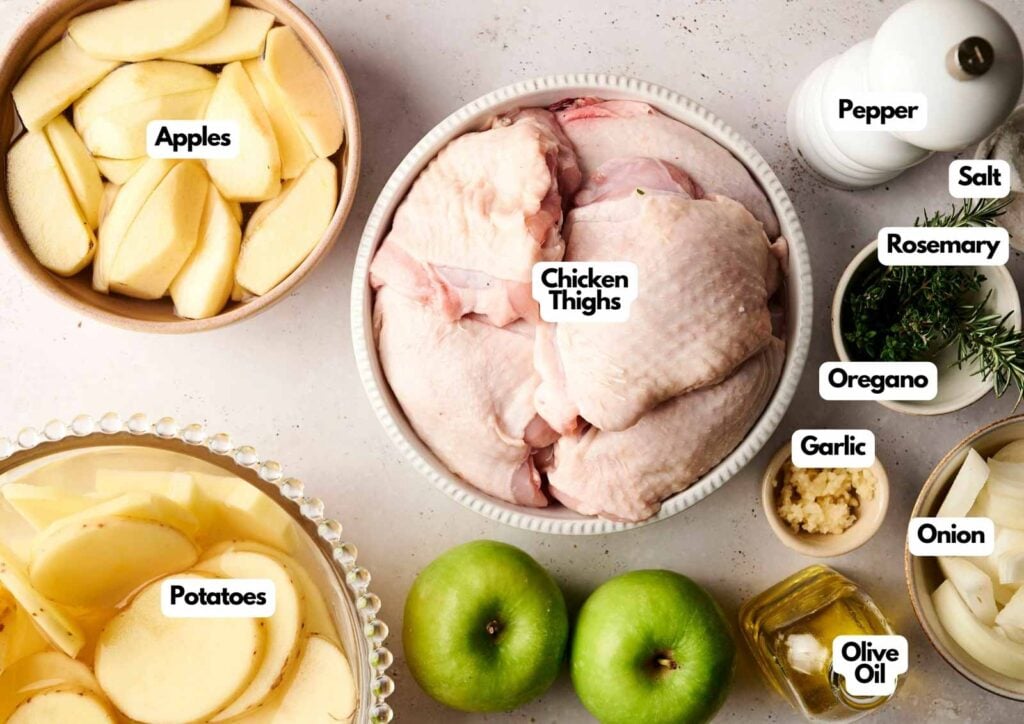 Sliced apples, potatoes, green apples, chicken thighs, chopped garlic, onion, olive oil, rosemary, oregano, salt, and pepper arranged on a countertop—perfect for making flavorful baked chicken thighs.
