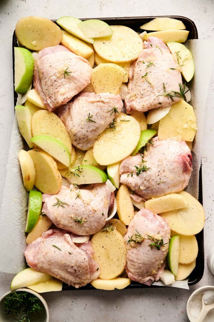 Baked chicken thighs, sliced potatoes, and apple slices are arranged on a sheet pan with sprigs of rosemary, ready to be cooked.