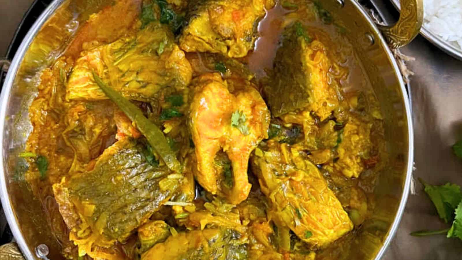 A bowl of Bengali Fish Curry with chunks of meat and vegetables, garnished with cilantro, served next to a plate of rice.